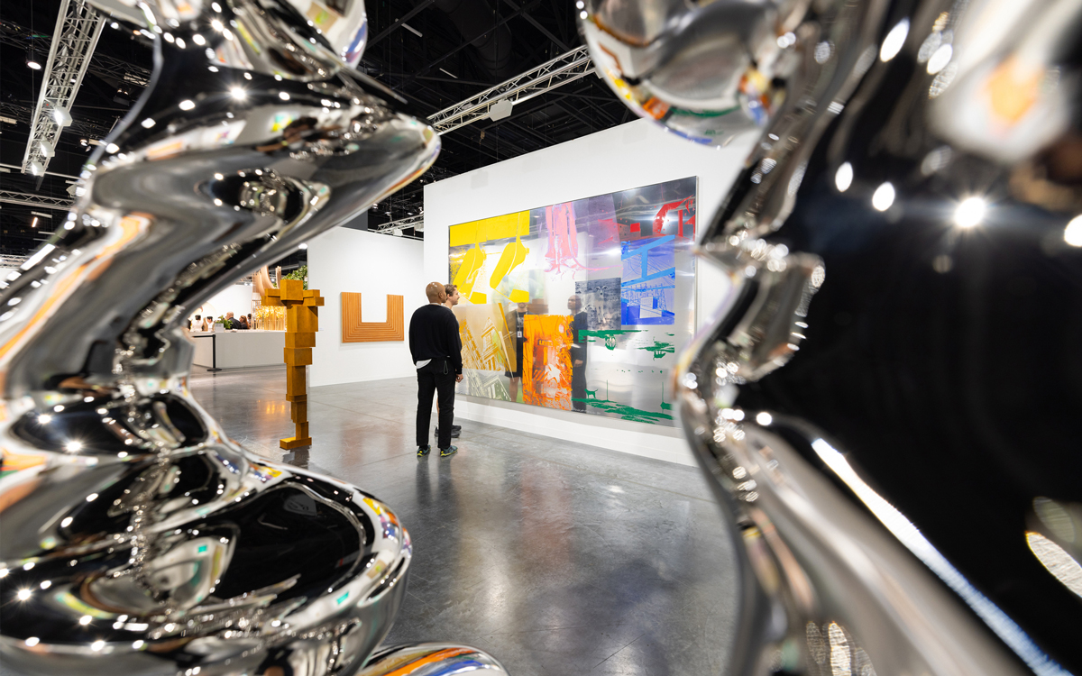 A view of an art fair through a stainless sculpture. Two men look at a painting.