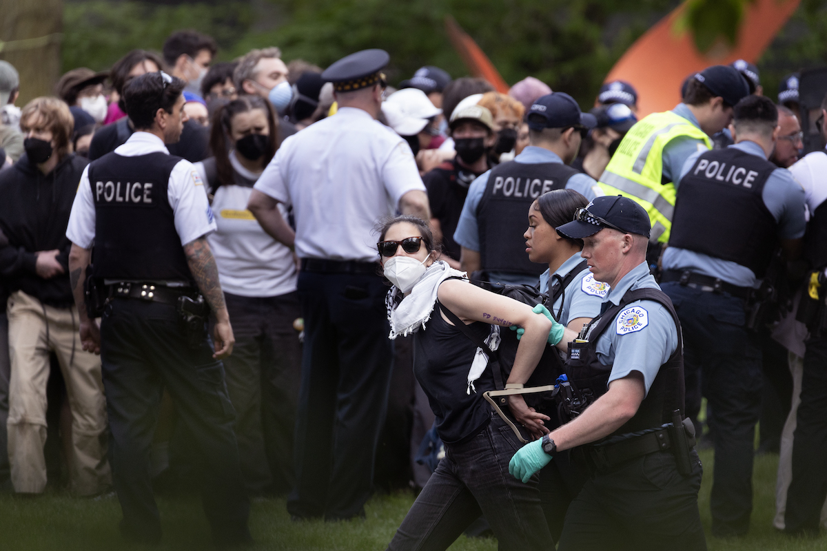 A person in a face mask and sunglasses being marched away by two police officers. Behind them, there are four other police officers and other protestors.