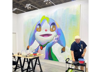 An artist stands in front of a painting in progress at an fair. It shows a close-up of a smiling animal-like figure who has one hand raised.