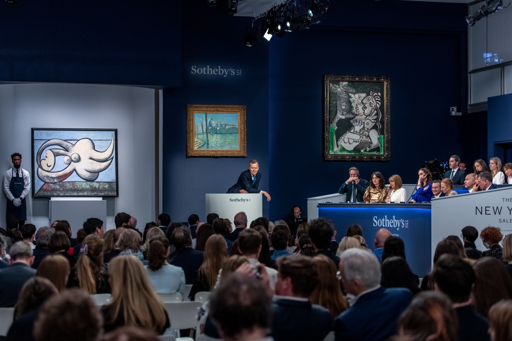 Audience in auction room with paintings.